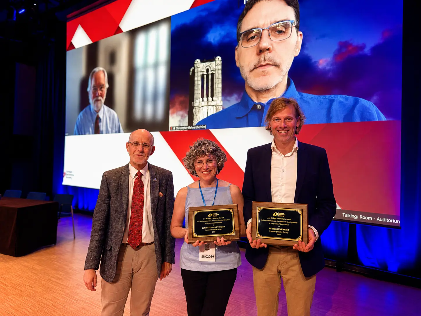 Three people stand on stage with two people on a screen behind them.