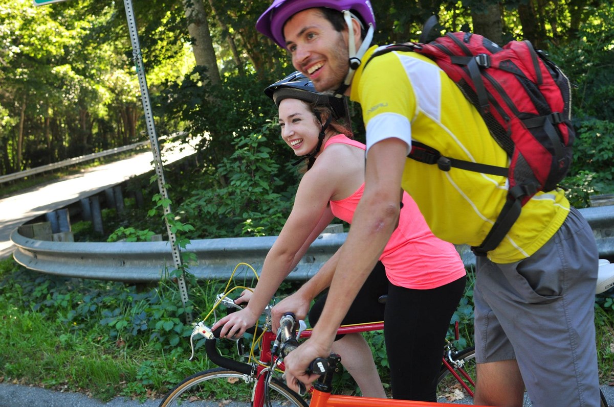 Two people wearing bike helmets and a backpack and riding bikes down a road with a guardrail and woods.
