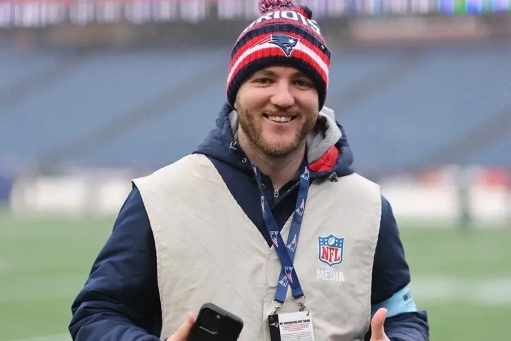 A young bearded man smiles for a photo on a football field while wearing a Patriots stocking cap and an NFL media vest. 
