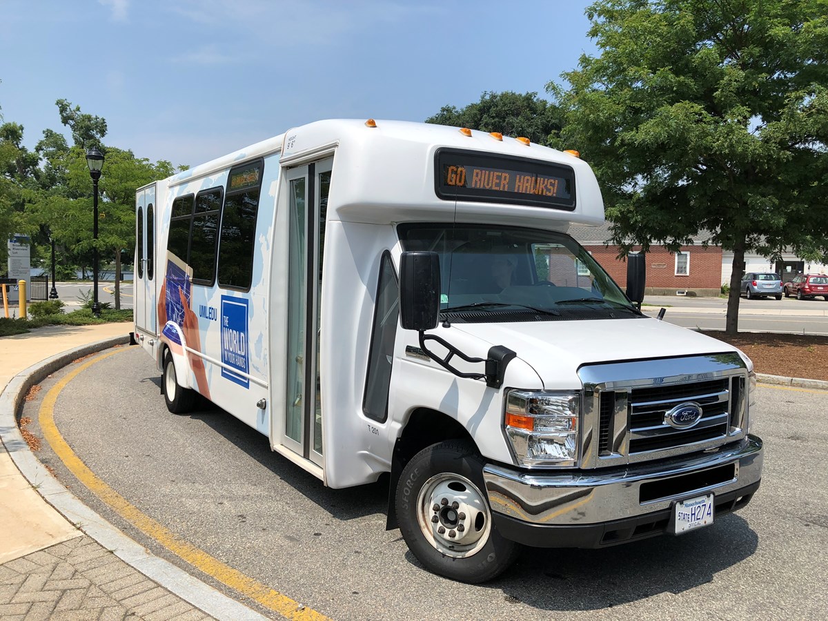 A white UMass Lowell shuttle stopped at a bus stop on South Campus. The LED sign in the front reads "Go Riverhawks" 