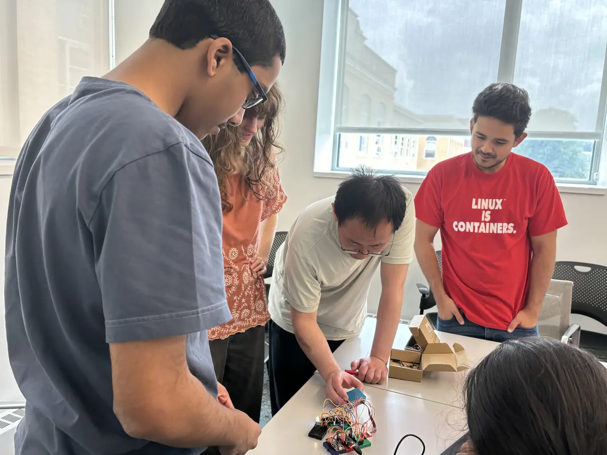 An instructor inspects an electronic device and wiring as a group of students look on