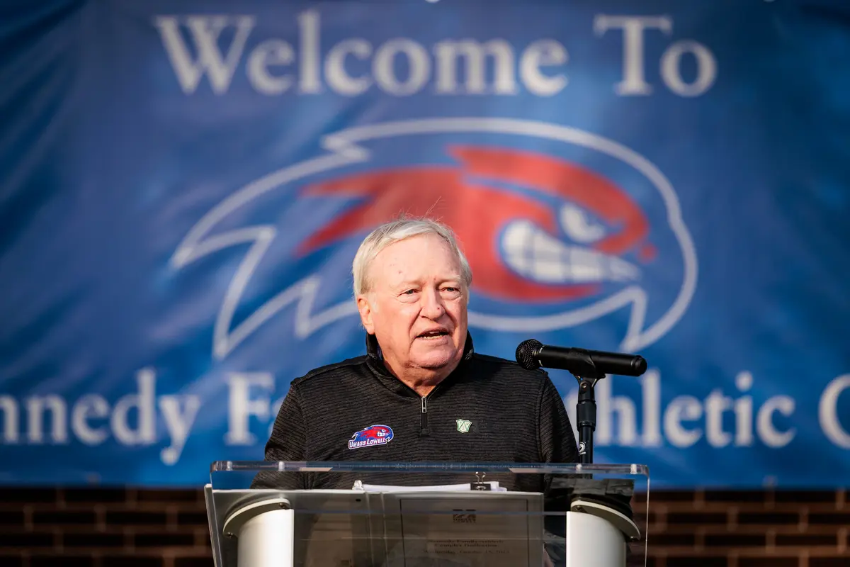 Man standing at a podium with a blue and red River Hawks banner hanging in the background