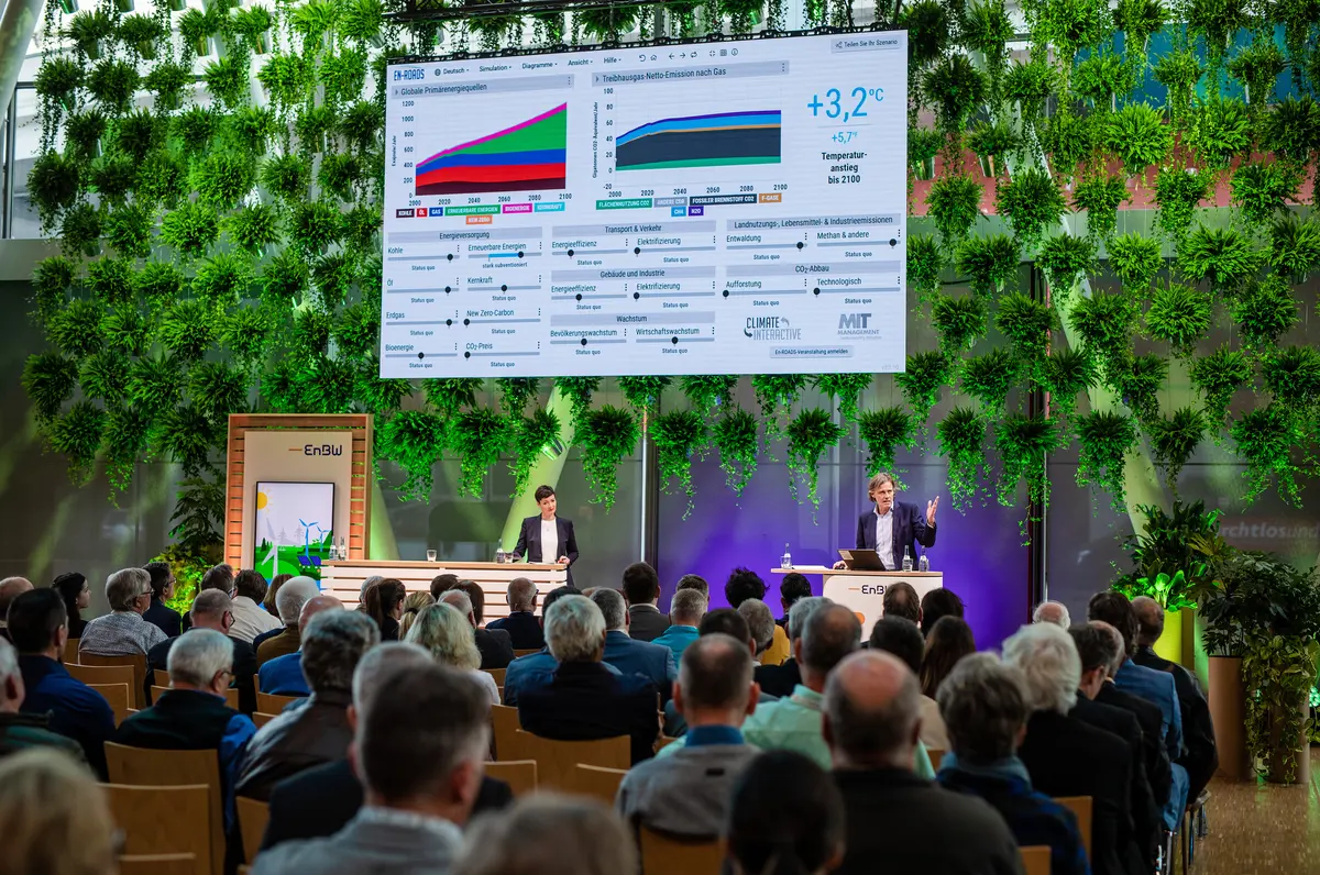 A screen showing a graphic dashboard with colored line graphs appears above the heads of participants at a conference 