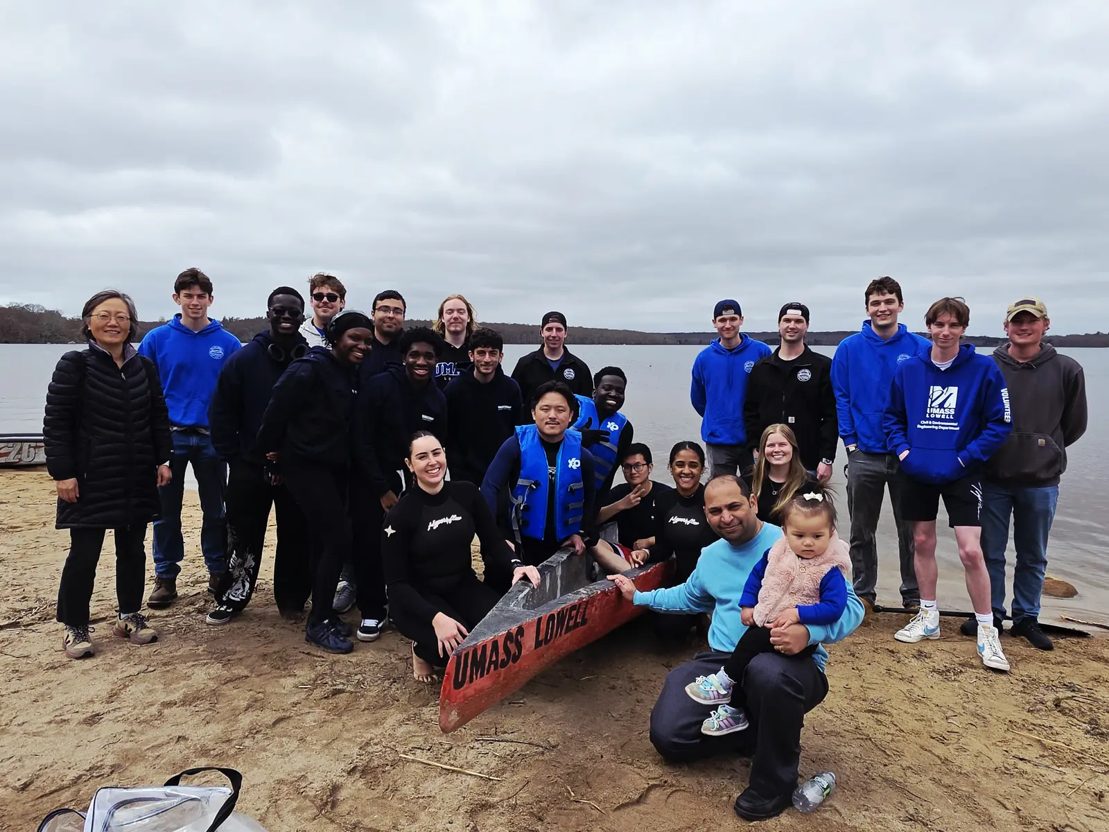 Two dozen people pose for a group photo alongside a concrete canoe on the shore of a lake.