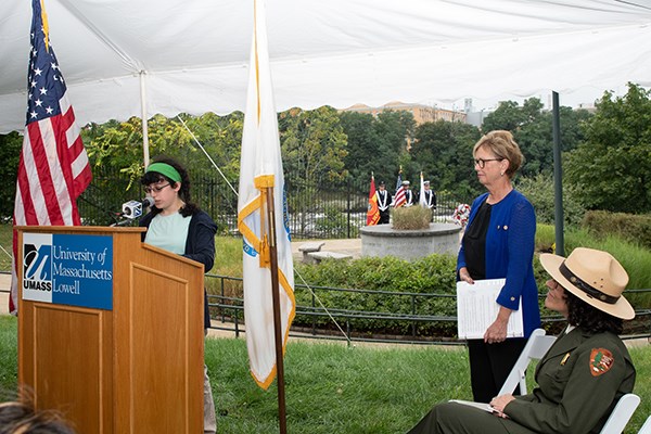 A student reads at a podium while Chancellor Moloney looks on in front of a 9-11 memorial