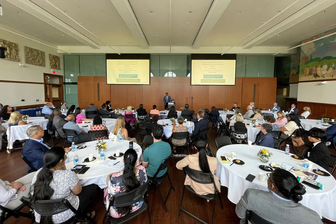 A person makes a presentation to a roomful of people seated at round tables.