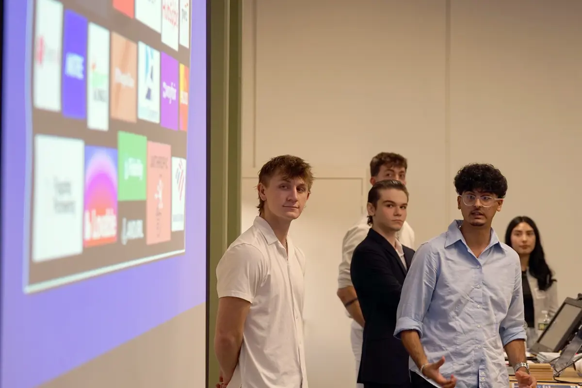 A group of students look at a projection screen while giving a presentation at the front of a room.