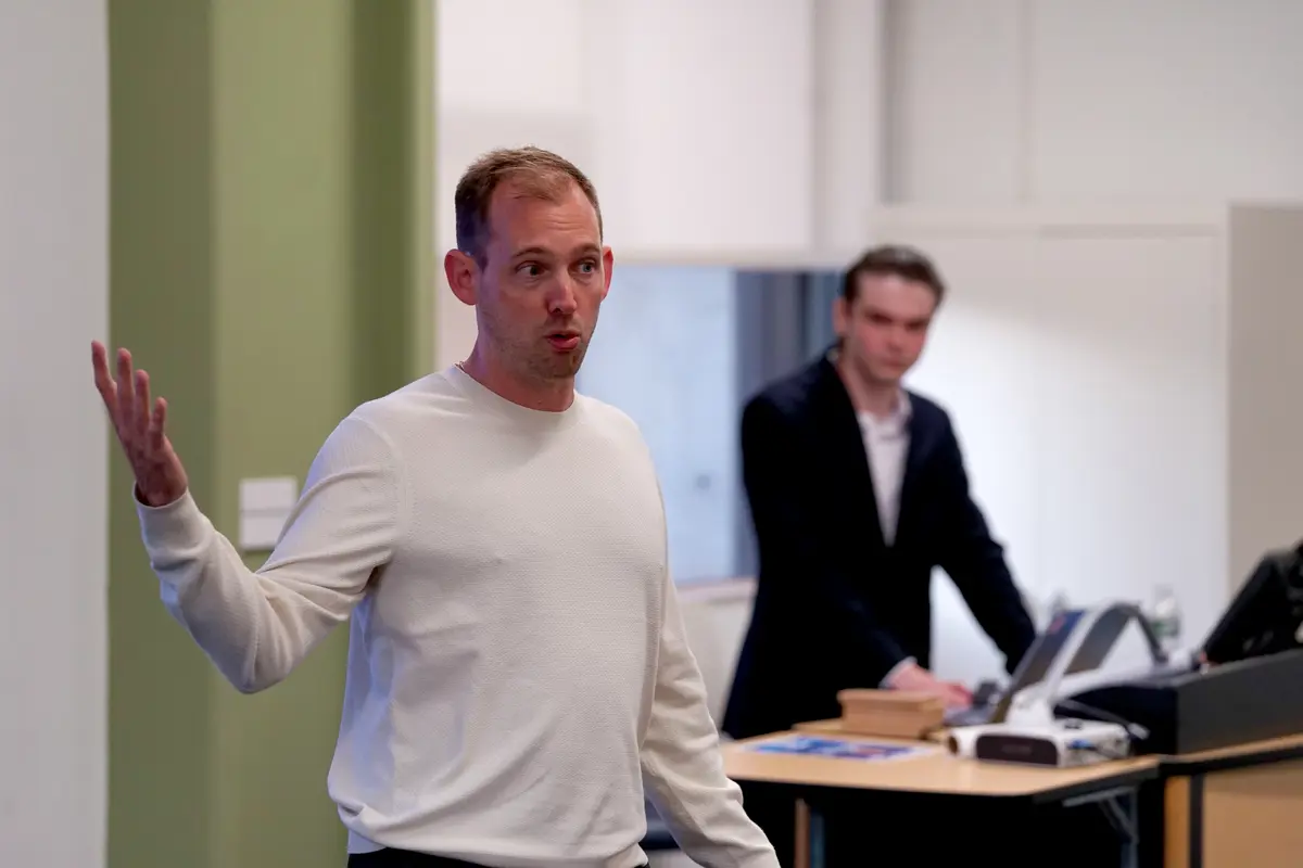 A man gestures with his hand while speaking at the front of a room as a person looks on in the background.