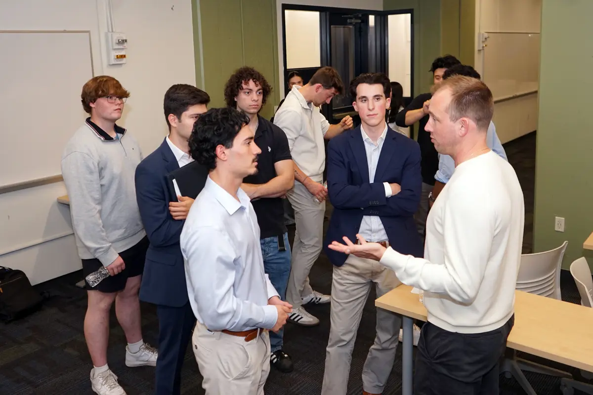 A man in a light colored seater talks to a group of college students.