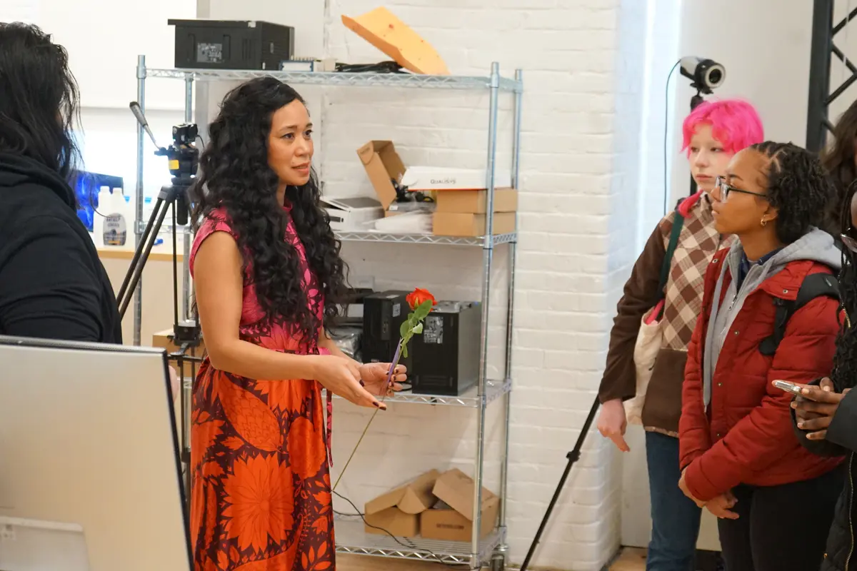 A woman in an orange dress talks to two students in a robotics lab.