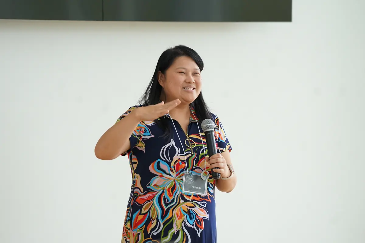 A woman gestures with her right hand and smiles while speaking into a handheld microphone.
