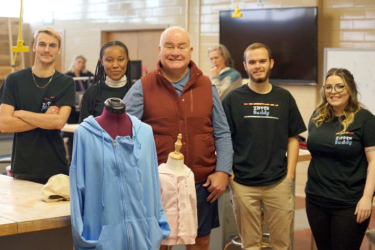 Four students wearing black T-shirts pose for a photo with a man wearing a brown vest. Two manequins are set up in front of them.