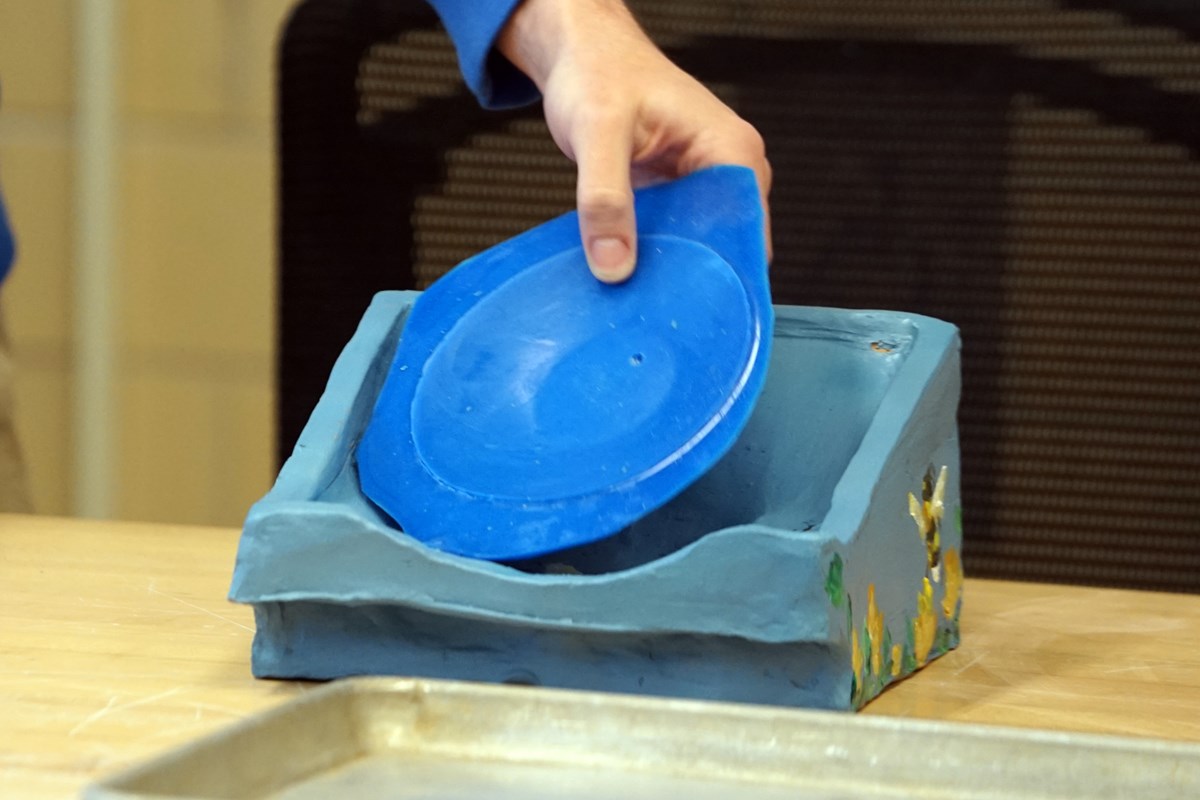 A hand is seen holding a blue piece of plastic over a ceramic stand, which has a bee painted on the side.