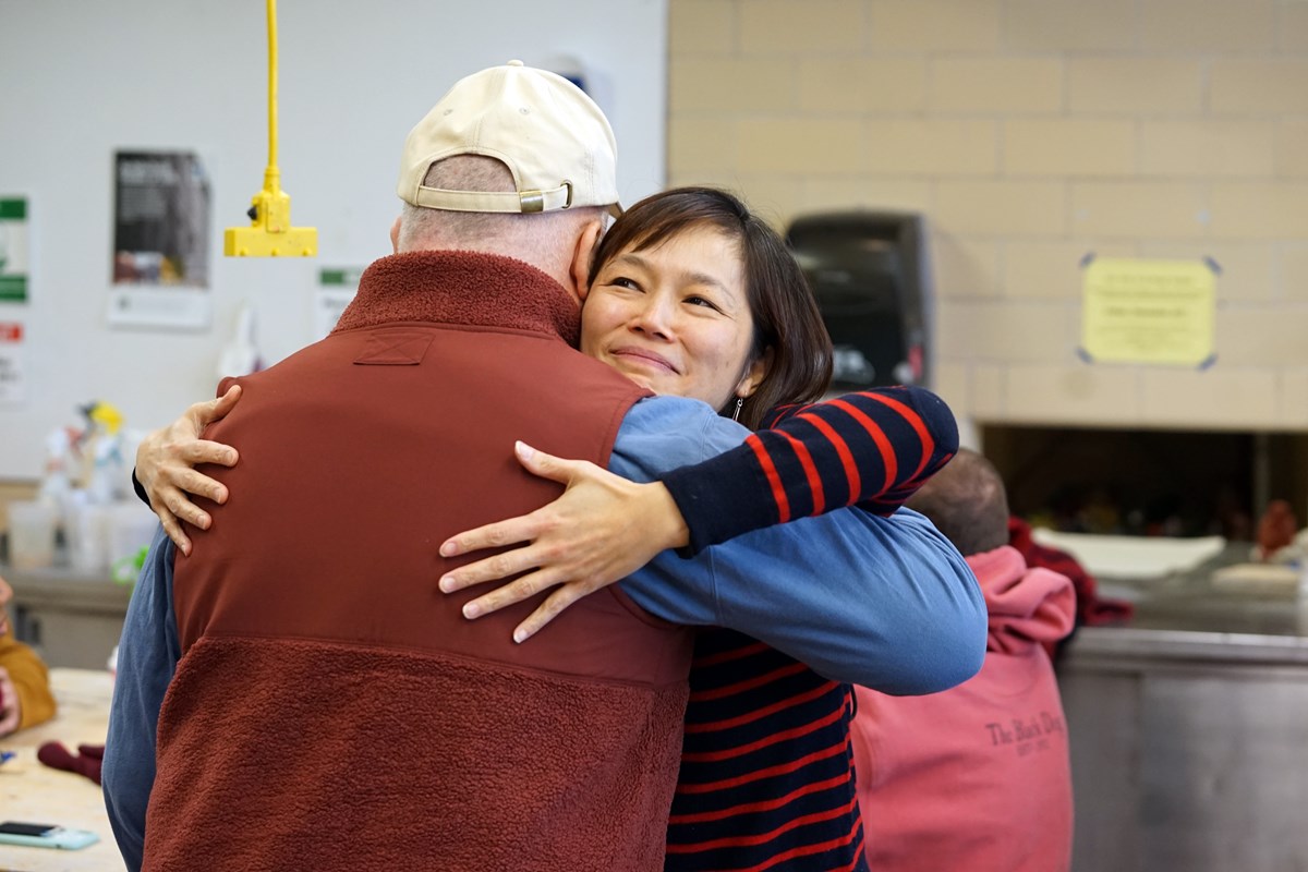 A woman in a black and red striped shirt hugs a man in a brown vest