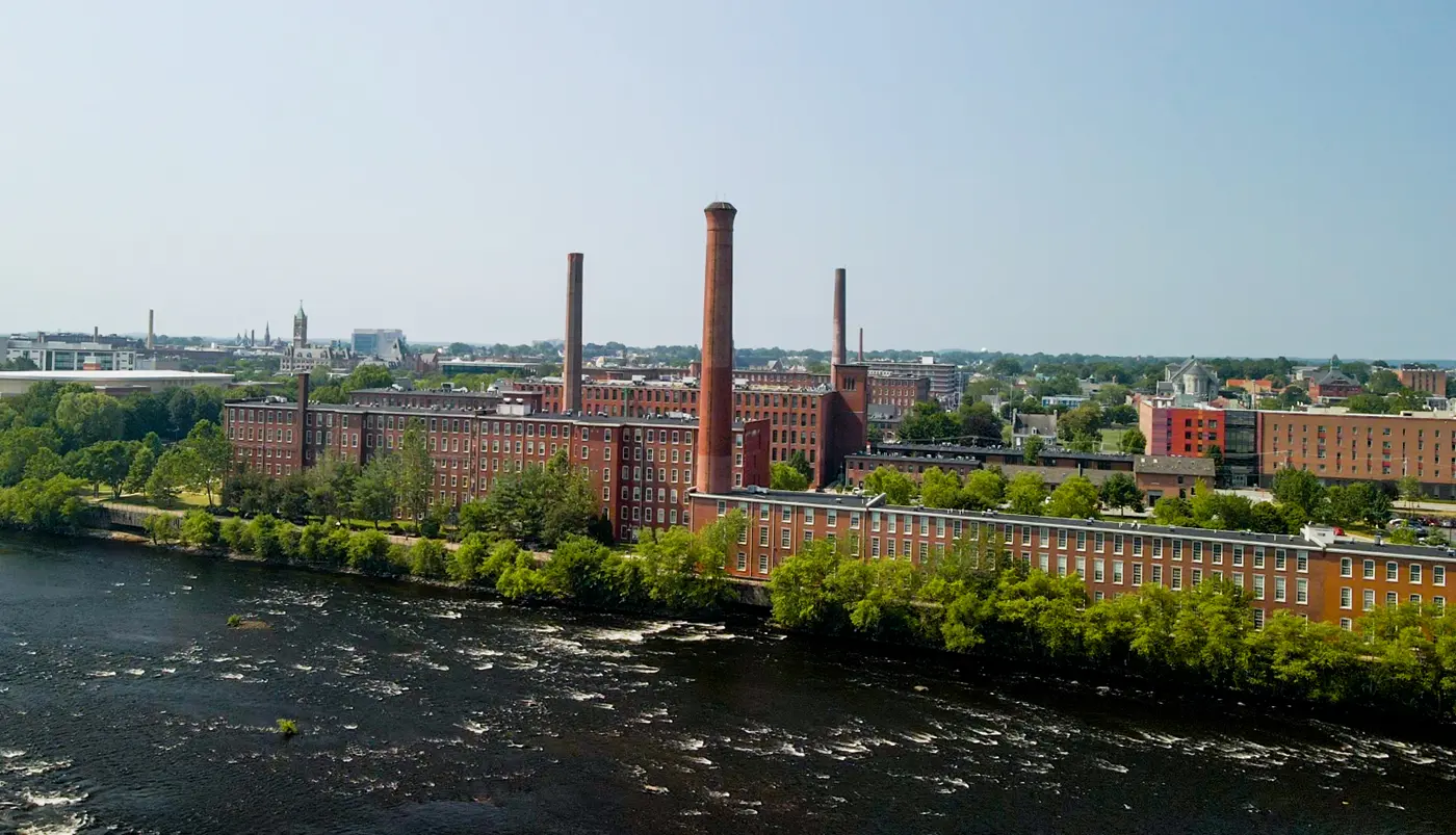 River flowing in foreground with trees alongside of massive red brick mill with smokestacks and city in distance