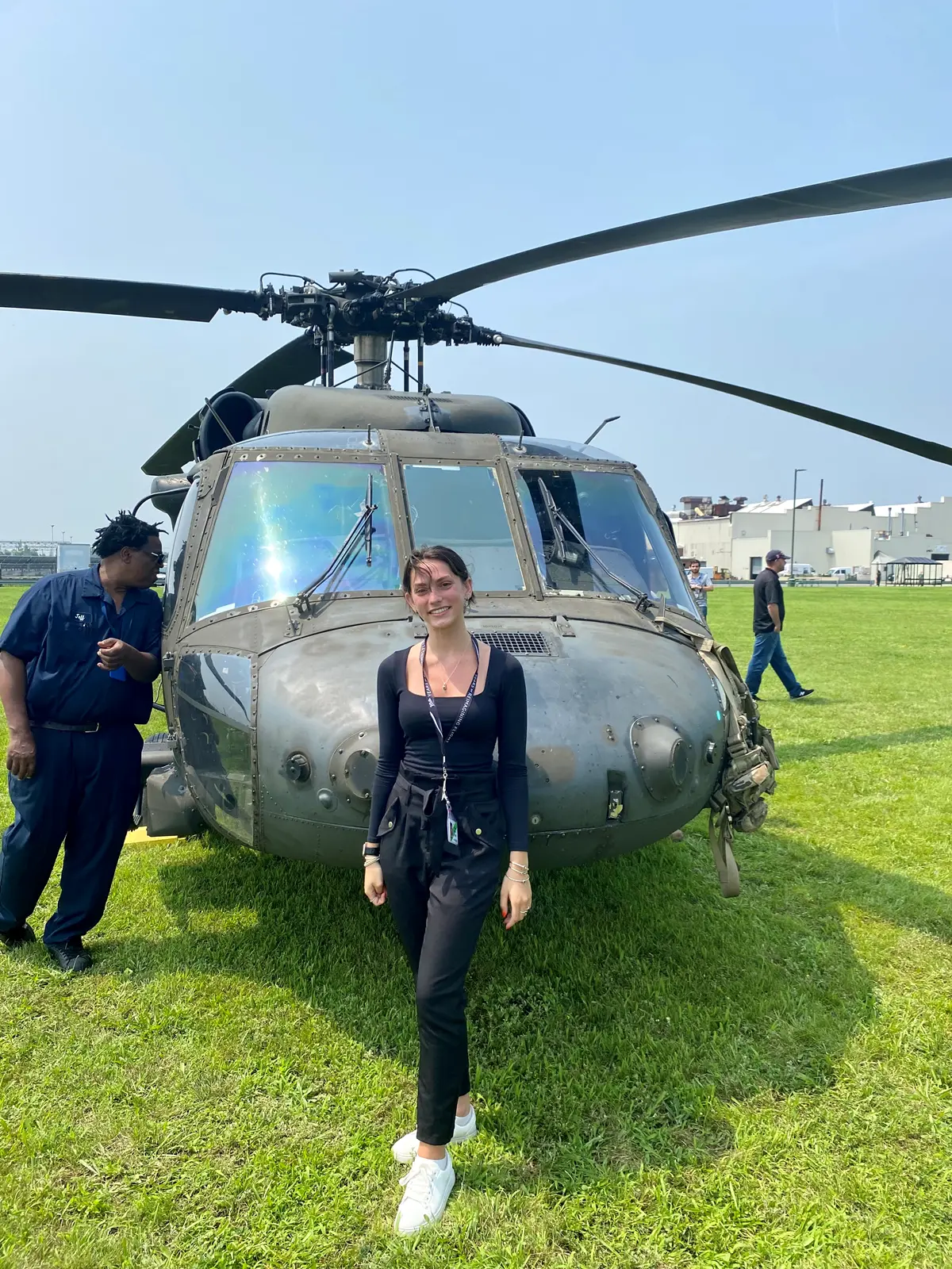 A young woman in a dark outfit poses for a photo in front of a military helicopter.