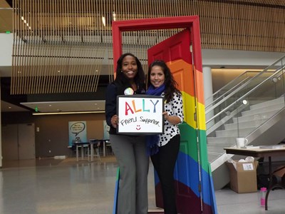 Two people standing in a rainbow-colored door, holding sign stating "Ally, friend, supporter"