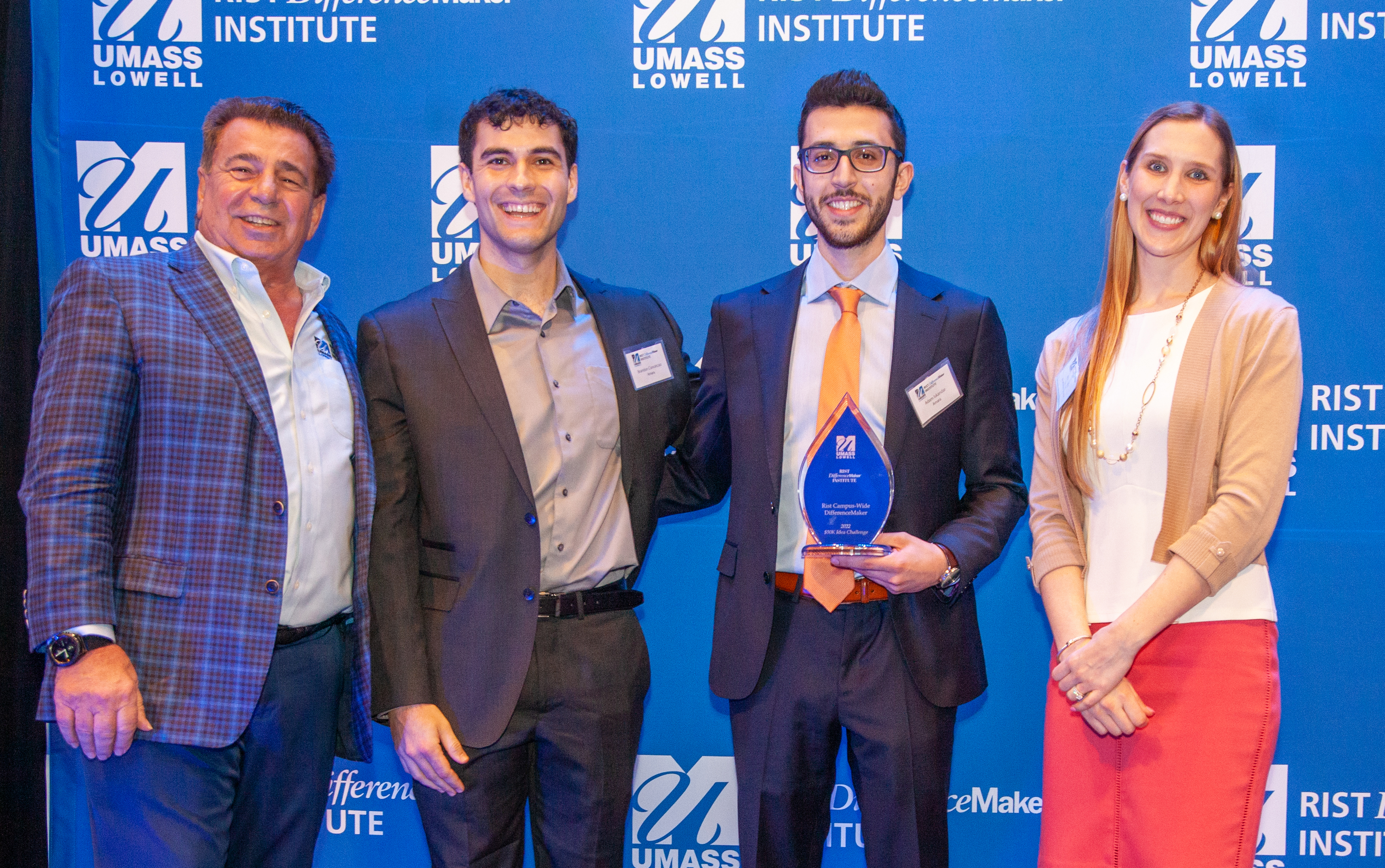 2 male students from the AMARA team holding an award pose with Brian Rist and Holly Lalos of Difference Makers against a blue UMass Lowell backdrop.