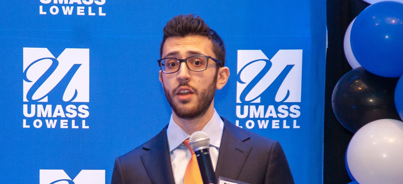 One of the members AMARA team holding a microphone and speaking in front of blue UMass Lowell backdrop.