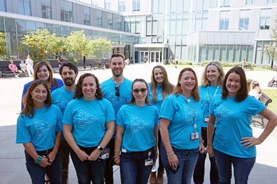 A group of men and women in blue T-shirts pose for a photo outside in front of a glass building