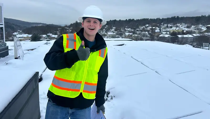 Andrew Fenner gives a thumbs up while standing on a roof and wearing a construction hat and vest.