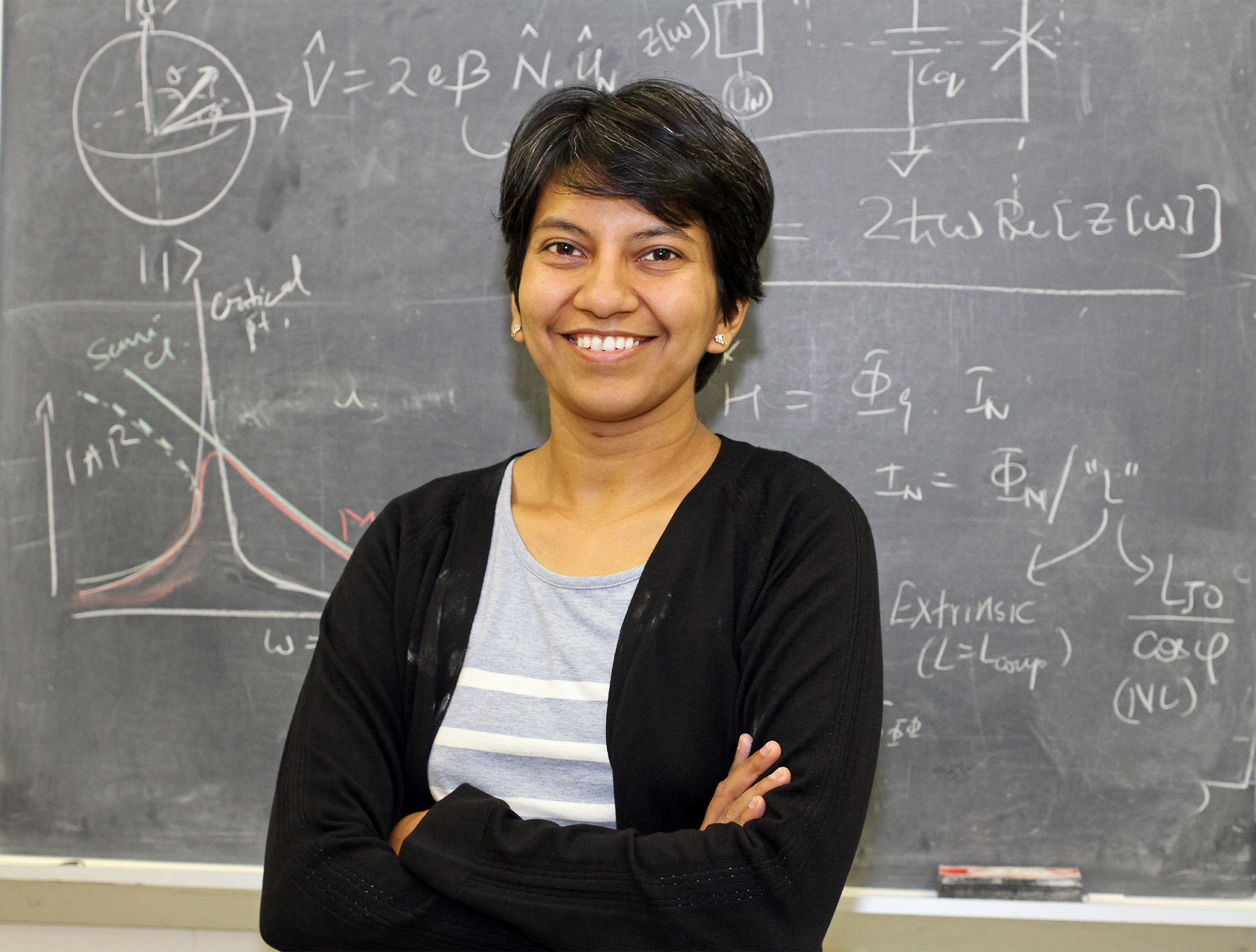 Physics Professor Archana Kamal smiling in front of a chalk board with writing and illustrations on it.