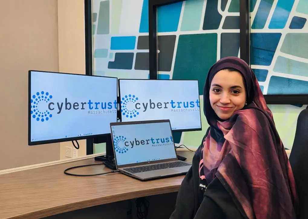 Asmaa Amiar sitting at a desk with a computer and monitors and the cybertrust logo.