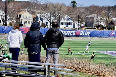 Three people stand and watch a women's soccer game on the field below