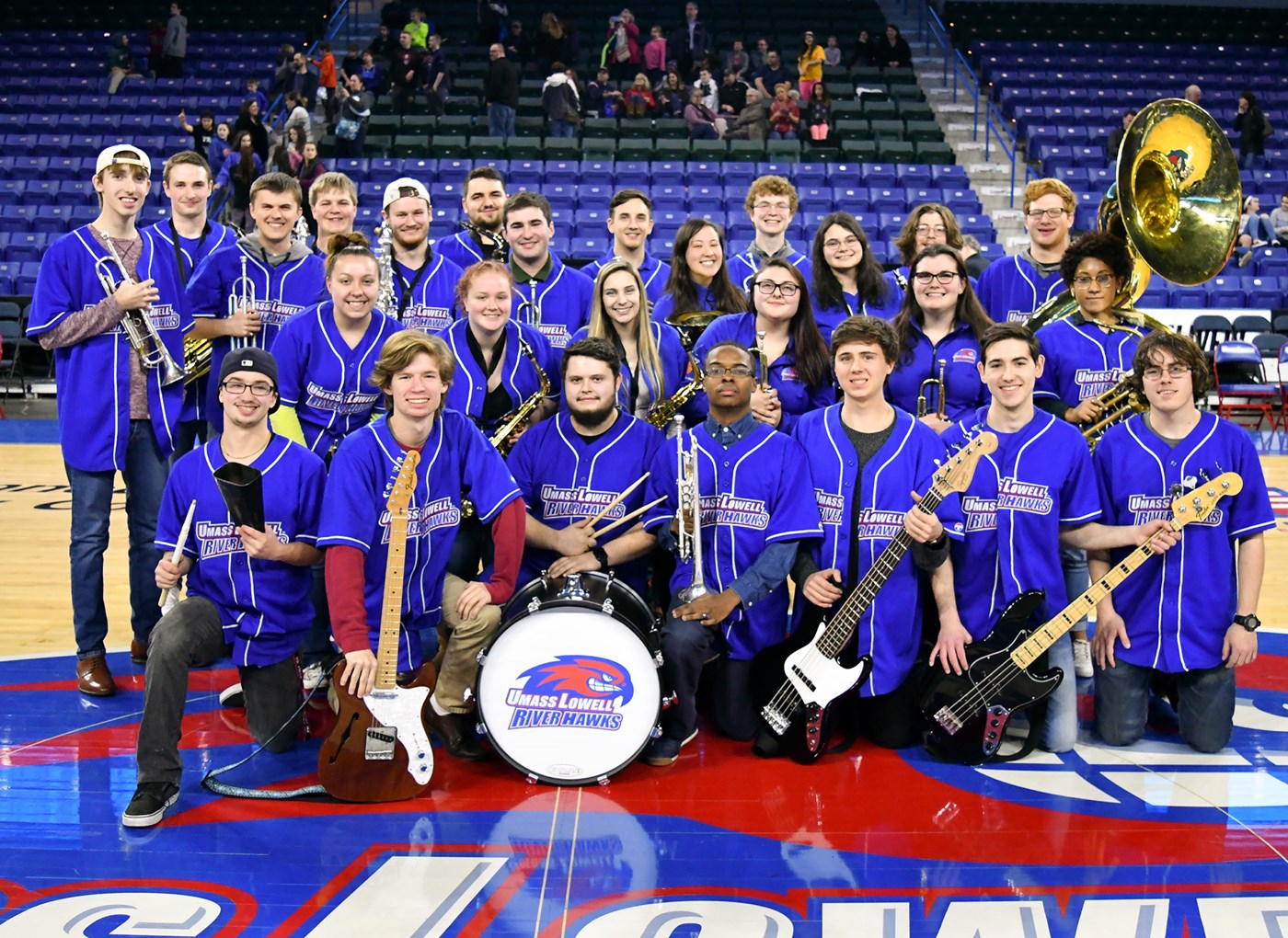  The UMass Lowell Basketball Pep Band is posed on center court wearing their royal blue short sleeved River Hawks Pep band shirts, with woodwind, brass and rhythm instruments like guitar and drums in hand.