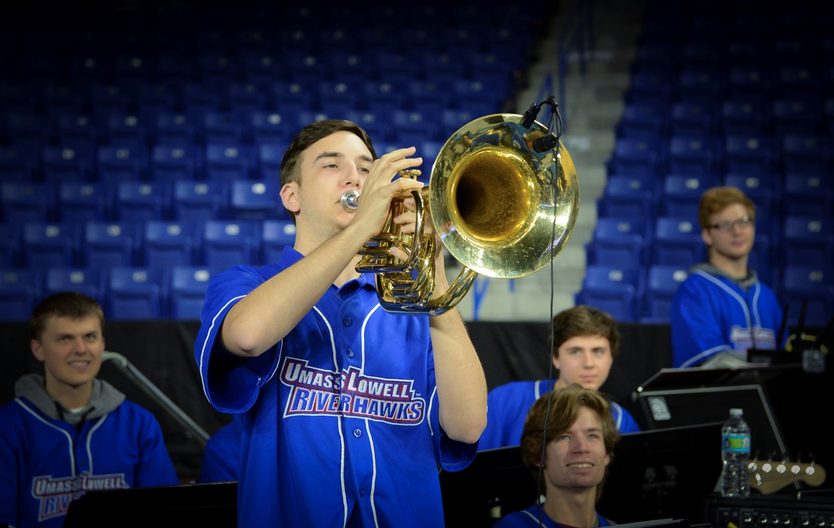 A UMass Lowell Basketball Pep Band baritone horn musician is featured standing, playing on a microphone, while band members holding trumpets and electric bass accompany him or wait for their turn.