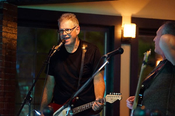 A man in glasses and a black T shirt sings into a microphone and plays guitar