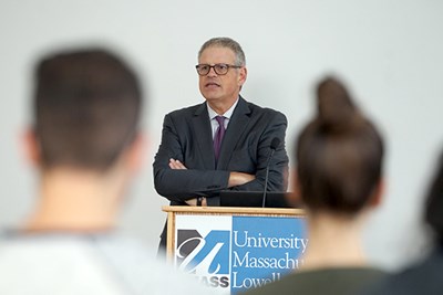 A man in glasses and a suit stands at a podium and talks to students