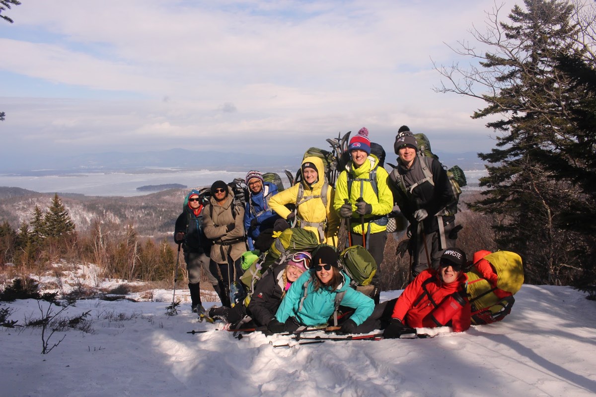 A group of happy hikers at the summit of the Mt. Major on the Belknap Range in the wintertime. The background of the photo shows a snow covered mountainside. 
