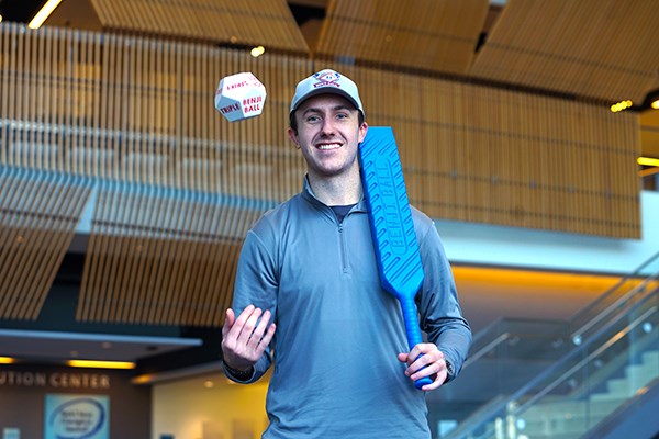 A young man in a baseball cap tosses a ball in the air while holding a blue plastic bat