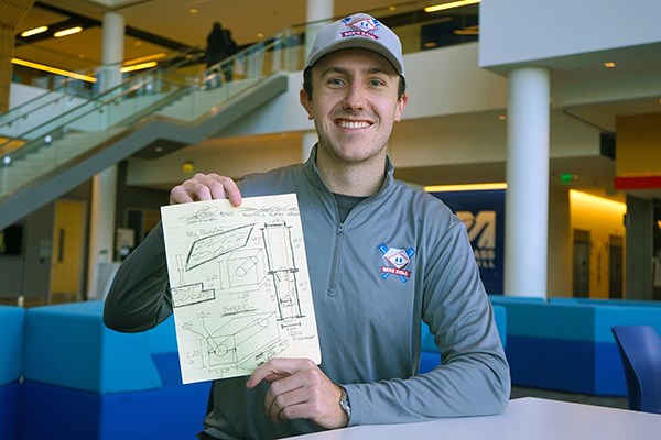 A young man in a baseball hat holds a piece of paper with drawings on it