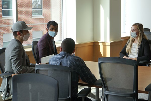 Four people wearing masks sit around a conference table in a room and talk