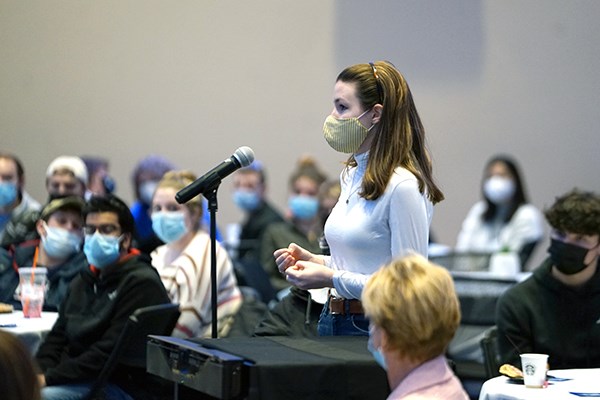 A young woman wearing a face covering stands at a microphone to ask a question while people in the room look on