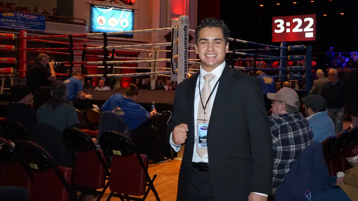 Brandon Diaz standing in front of a boxing ring with a clock on the wall and a crowd behind him.