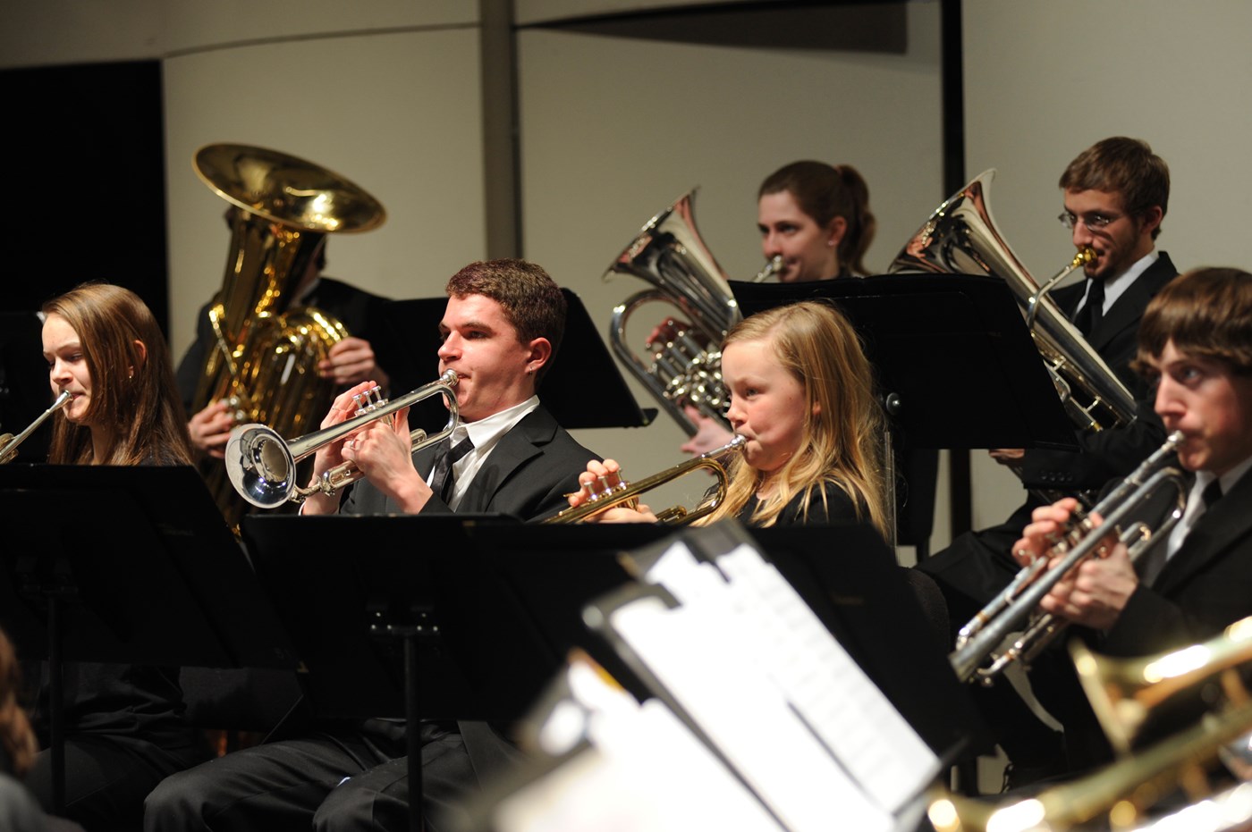 The Concert Band photo shows middle school trumpet and brass players seated next to college music performers also playing brass instruments each dressed in formal black concert dress performing music at Durgin Concert Hall on Umass Lowell’s South Campus.