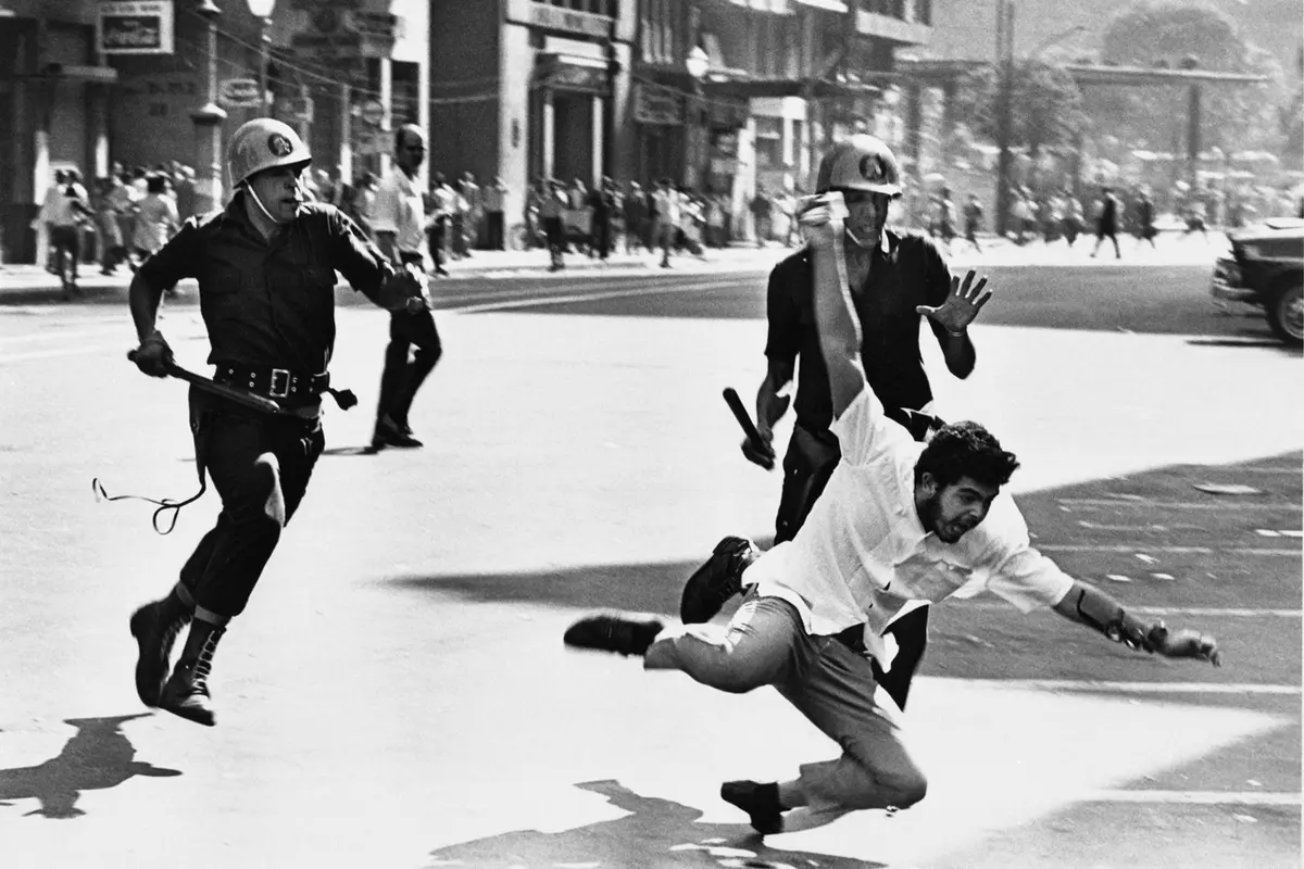 black and white photo of police with helmets and clubs chasing falling man