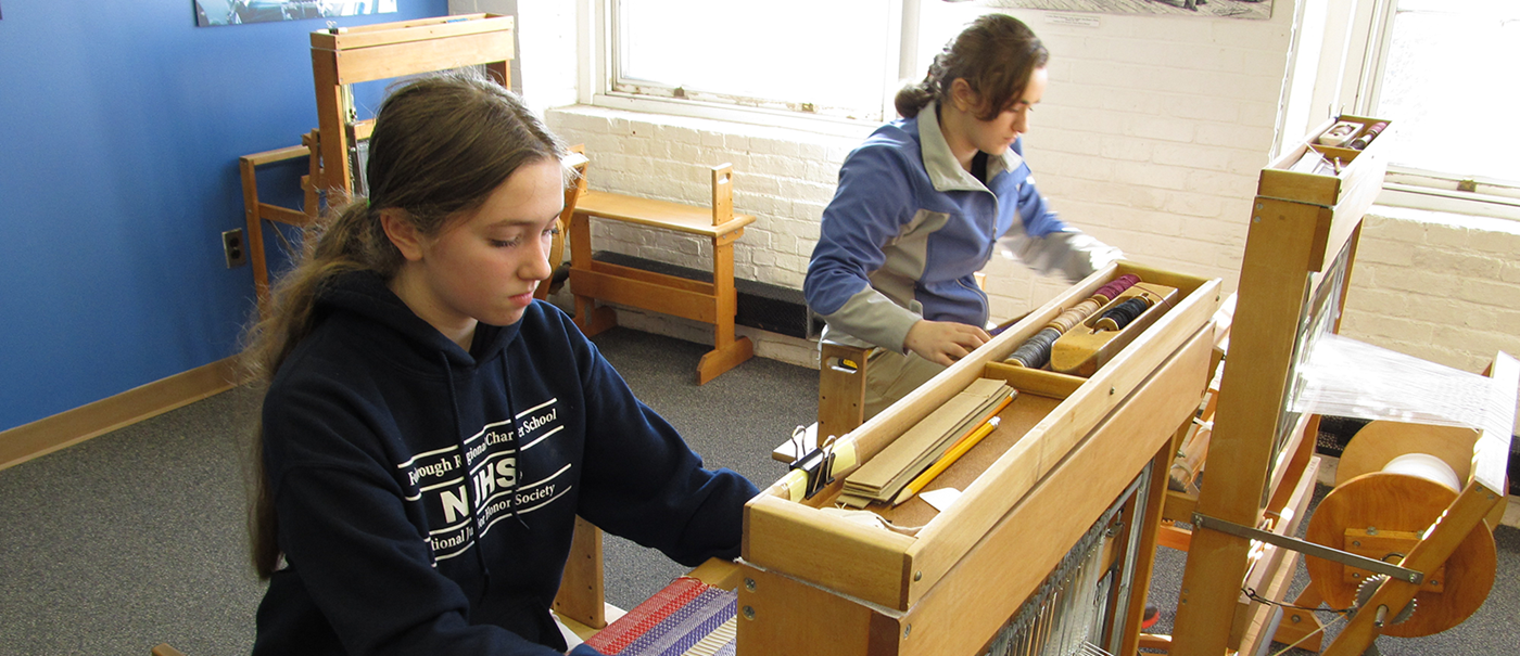 Two girls sitting in front of old looms weaving at Tsongas Industrial History Center