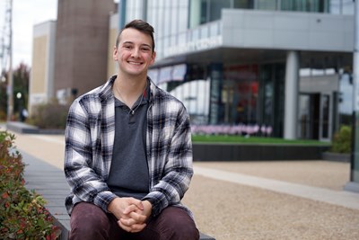 Jacob Villeneuve sitting outside and smiling in front of UMass Lowell's University Crossing. 