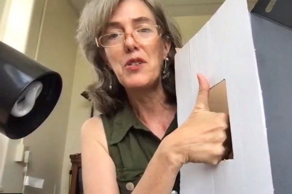 A woman in glasses holds a piece of cardboard that has a rectangular hole in the middle. Her right hand is in the opening.