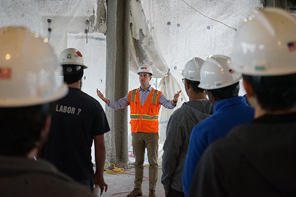 A man in an orange vest and hard hat gestures while talking to a group of students
