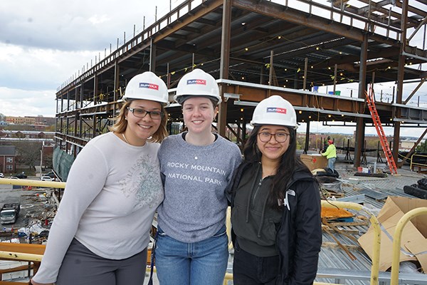 Three young women in white hard hats pose for a photo on a construction site