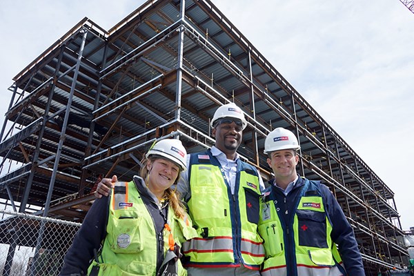 Two men and a woman in construction vests and hard hats pose for a photo in front of an unfinished building