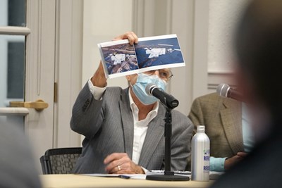 A man in a mask holds up a pamphlet while talking 