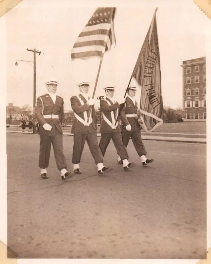 4 cadets marching down the street with American and Lowell Textile Institute flags.