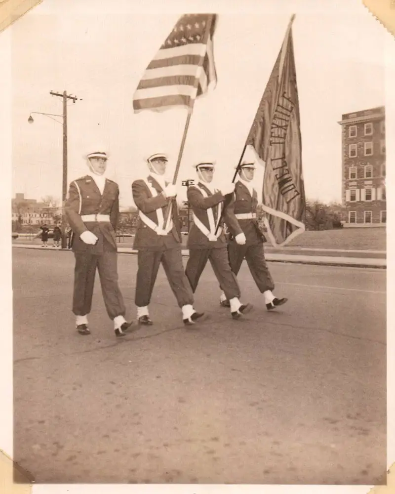 4 cadets marching down the street with American and Lowell Textile Institute flags.