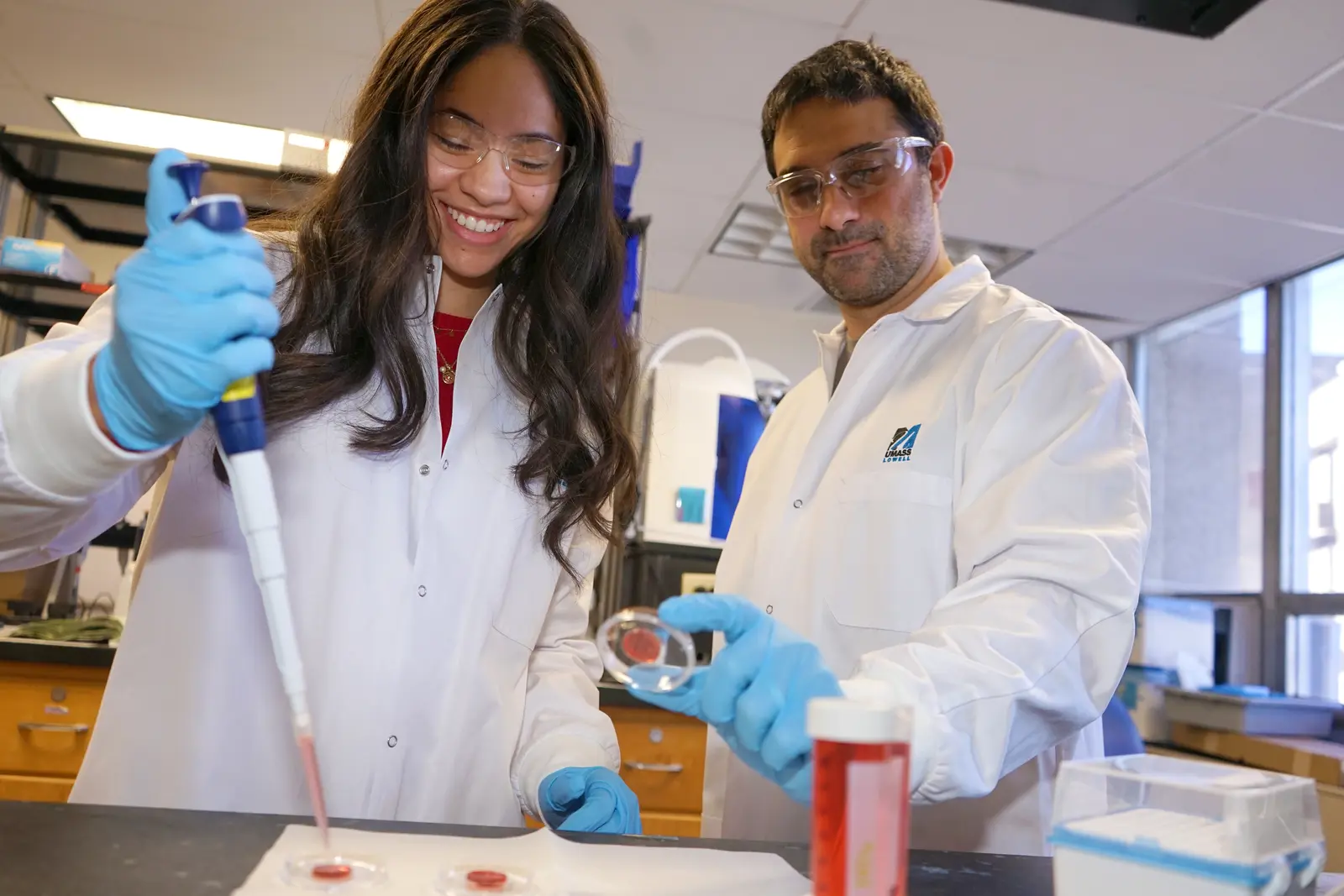 A young woman in a lab coat pipettes a specimin while a man in a lab coat holds up a petri dish.